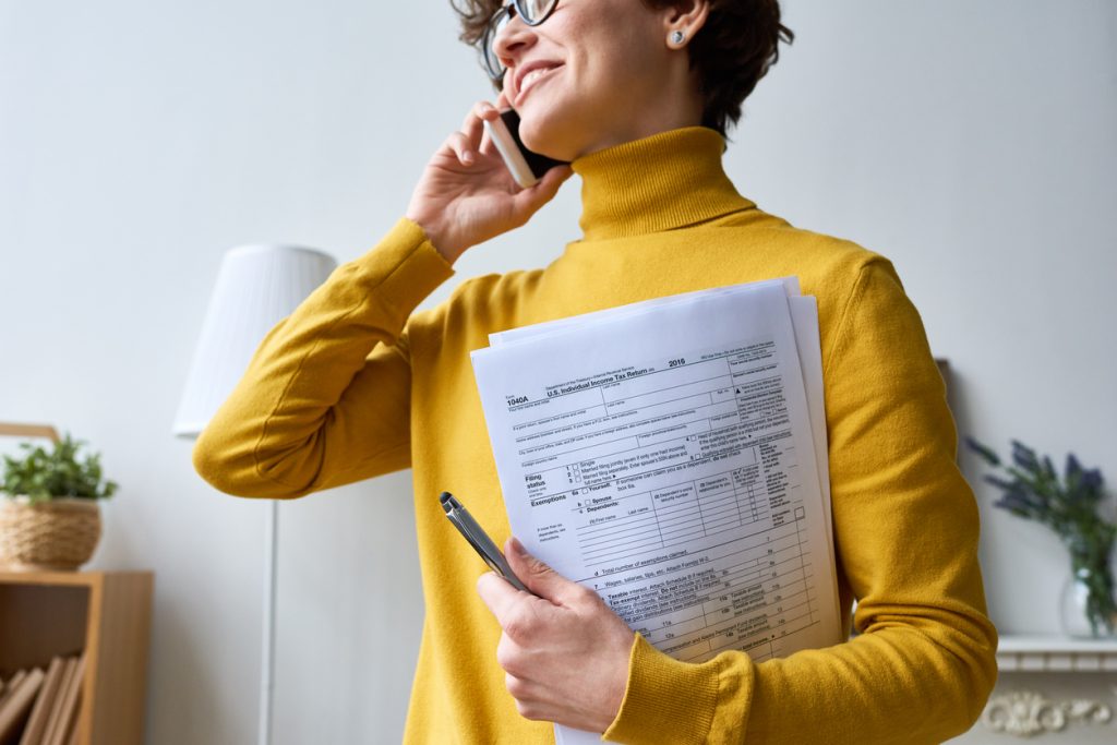 Cheerful woman in yellow sweater calling on phone and holding income tax return papers